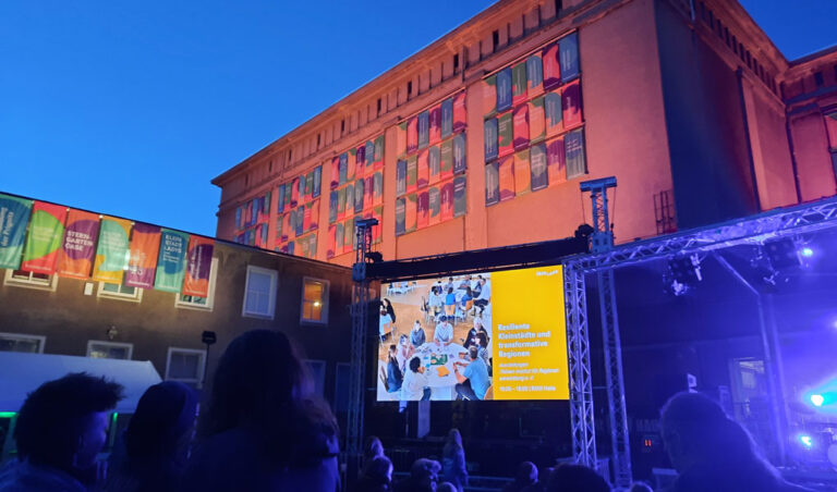 Das Kühlhaus Görlitz am Abend beim Überlandfestival 2025. Viele farbige Fahnen am Gebäude zeigen preisgekrönte Initiativen.