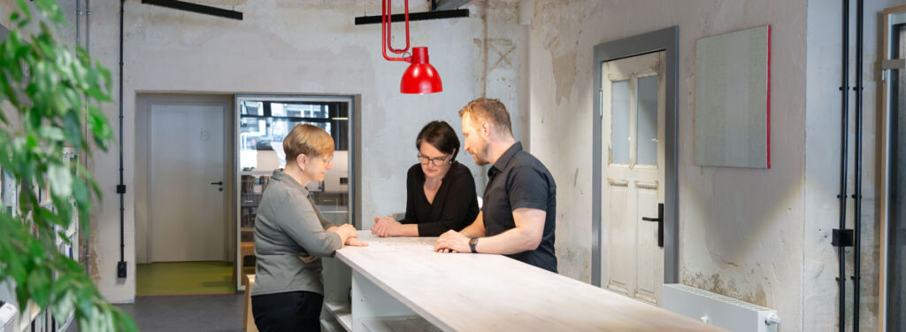 Drei Personen stehen an einem Tresenmöbel und schauen gemeinsam auf ein Dokument. Über ihnen eine rote Leuchte. Links ragt eine Pflanze ins Bild. Ein Beispiel für agile Arebitswelten mit spontanen Stehbesprechungen.