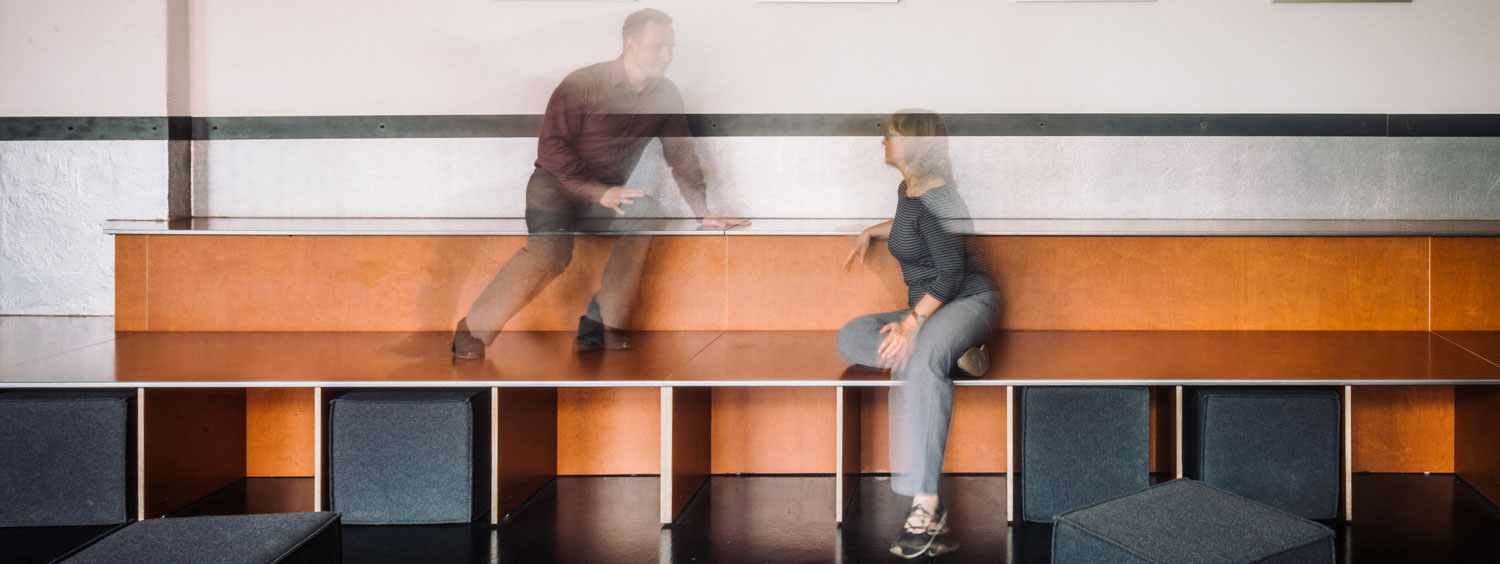 Zwei verwischte Personensilhoutten sitzen auf einer orangebraunen Stufeninstallation aus Holzwerkstoff. Im Vordergrund sind anthrazitfarbene Polsterwürfel zu sehen, die auch unter der Bank verstaut sind. Ein Beispiel für eine andere Art der Zusammenkunft anstelle eines klassischen Besprechungsraumes. Beim Recruiting können alternative Räume Vorteile bieten.