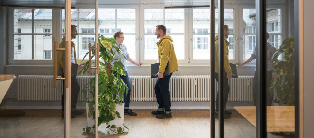 Zwei Personen stehen vor einem Fenster im spontanen Gespräch. Rechts und links stehen Telefonboxen als Zeichen einer modernen Arbeitswelt. Eine Grünpflanze belebt das Bild.