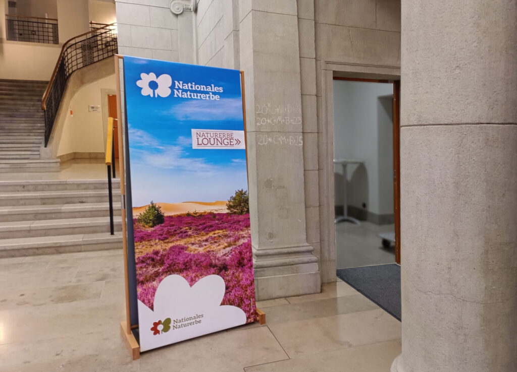 Ein großer Holzaufsteller mit einem Plakat in einem historischen Foyer mit Natursteintreppe und Säulen. Auf dem Plakat ist eine Heidelandschaft zu sehen und Logo und Schriftzug "Nationales Naturerbe".