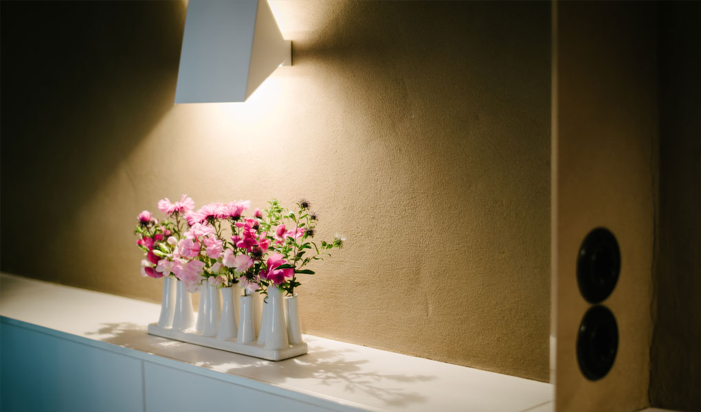 Nahaufnahem von Lehmputz in einem Bad. Man sieht in einem Spiegel eine weiße Wandleuchte und eine Blumenvase.