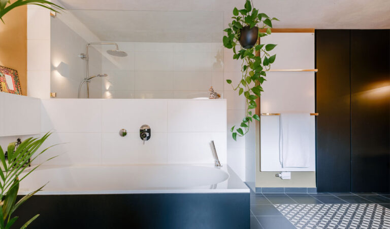 Ein Bad mit Badewanne, Dusche hinter einer Glastrennwand und ein Flächenheizkörper im Hintergrund. Farbgebung schwarz, weiß und etwas Lehmputz. Rechts unten im Bild Musterfliesen von Zahna-Fliesen.