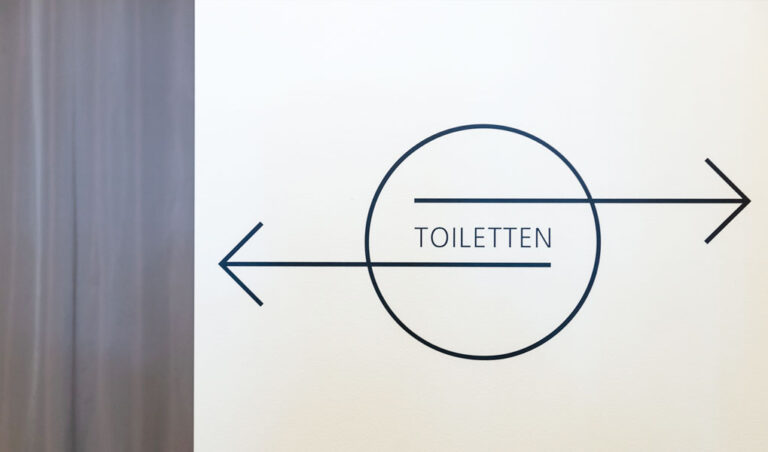Man sieht eine filigrane schwarze Grafik mit dem Wort Toiletten in einem Kreis mit Pfeilen nach rechts und links.