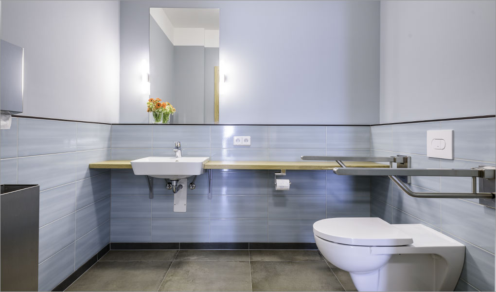Barrierefreies WC in einer Firma. Hellblaue Fliesen, hellblaue dezente Wandfarbe. Das unterfahrbare Einbauwaschbecken liegt auf einer schmalen Holzplatte.