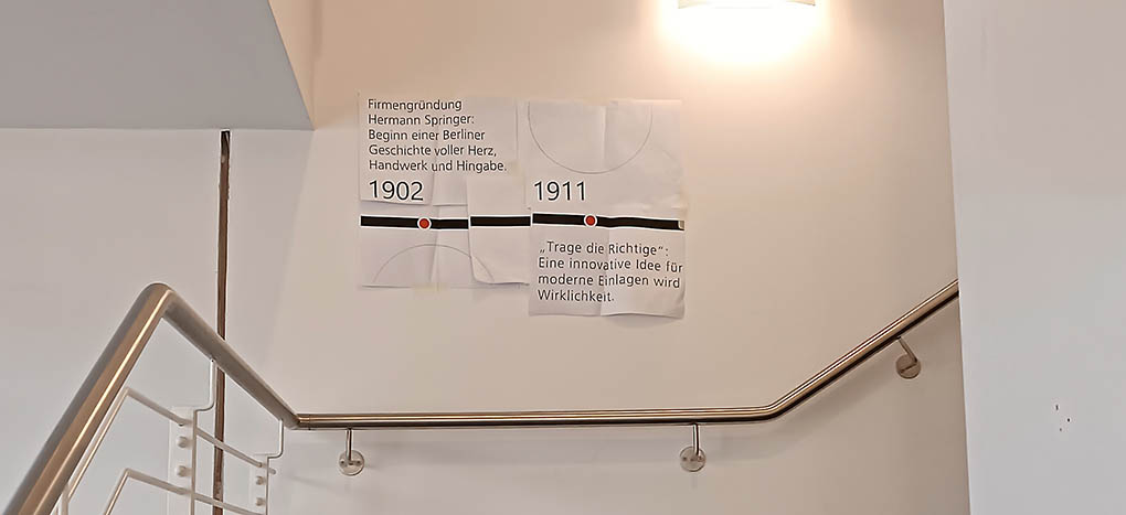 Über einem Treppengeländer aus Edelstahl hängen an einer weißen Wand Papierausdrucke von grafischen Linien und Text. Sie dienen als Probe-Ansicht für die schriftgröße von Wandbeklebungen innerhalb einer Ausstellungsplanung.