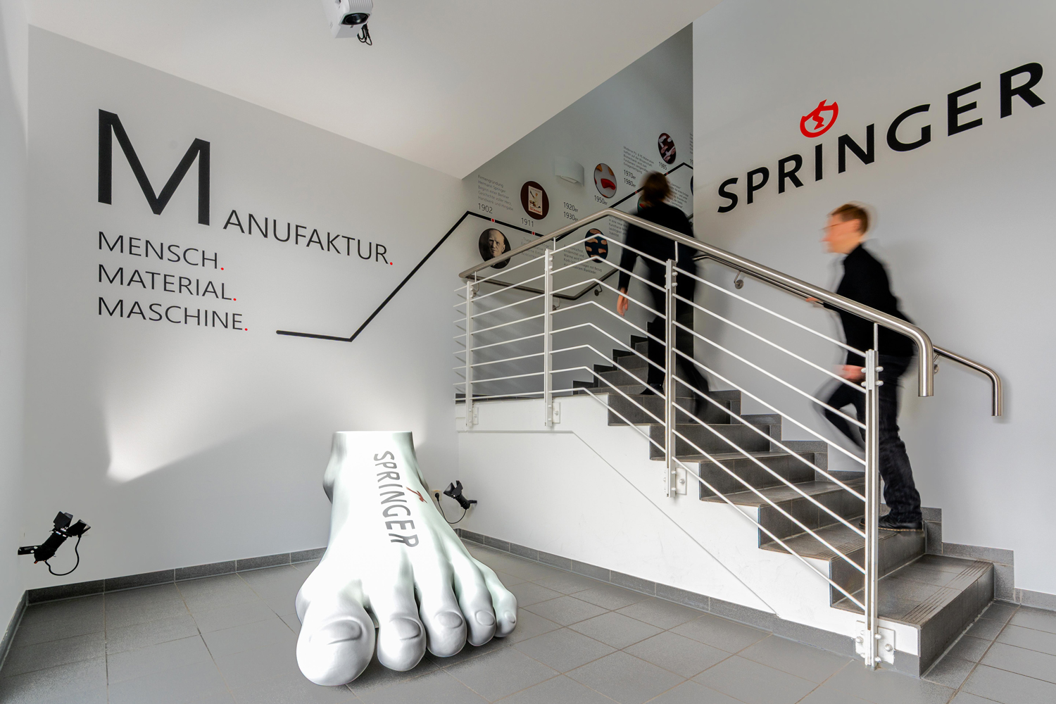 Ein kleines Foyer mit einem Treppenaufgang in einem pragmatischen Gewerbebau. Im Vordergrund eine überdimensionale Fußskulptur mit Logo-Projektion. Weiße Wände. An einer Wand das Logo der Springer Aktiv GmbH. An der anderen Wand die Schriftzüge "Manufaktur. Mensch. Maschine. Material". An der Wand des Treppenaufgangs ein Zeitstrahl zur Firmengeschichte mit kreisrunden Bildern und Text. Zwei Personen gehen die Treppe hoch.