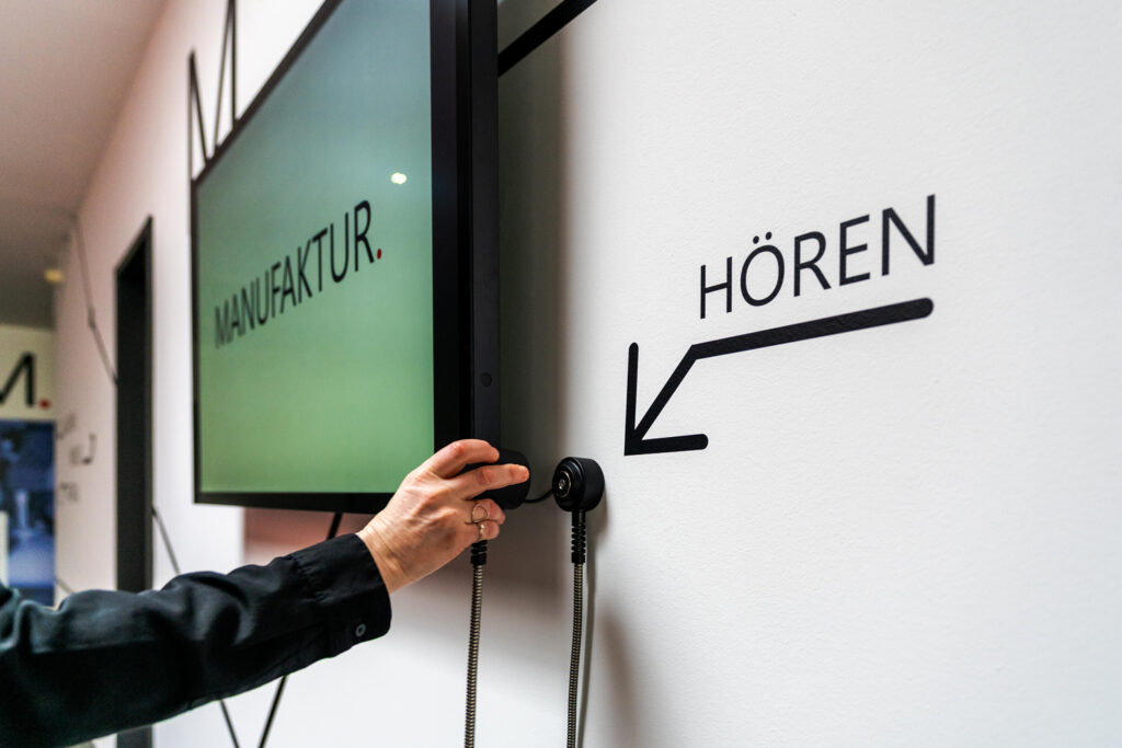 Nahaufnahme einer Hörstation neben einem Touchscreen. Auf der Wand sieht man das Wort "Hören" und einen Pfeil, der auf de Einhandhörer zeigt.