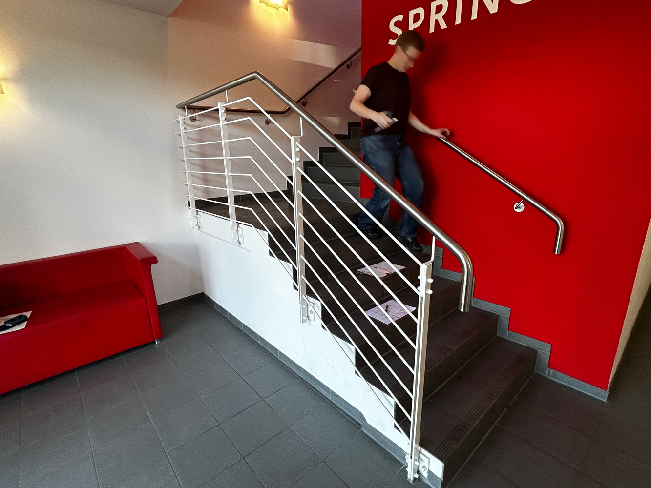 Vorherbild eines Foyers mit Treppenaufgang. Eine Wand ist strahlend rot und trägt das Logo der Springer Aktiv GmbH in weiß. In der linken Bildhälfte steht ein rotes Ledersofa.