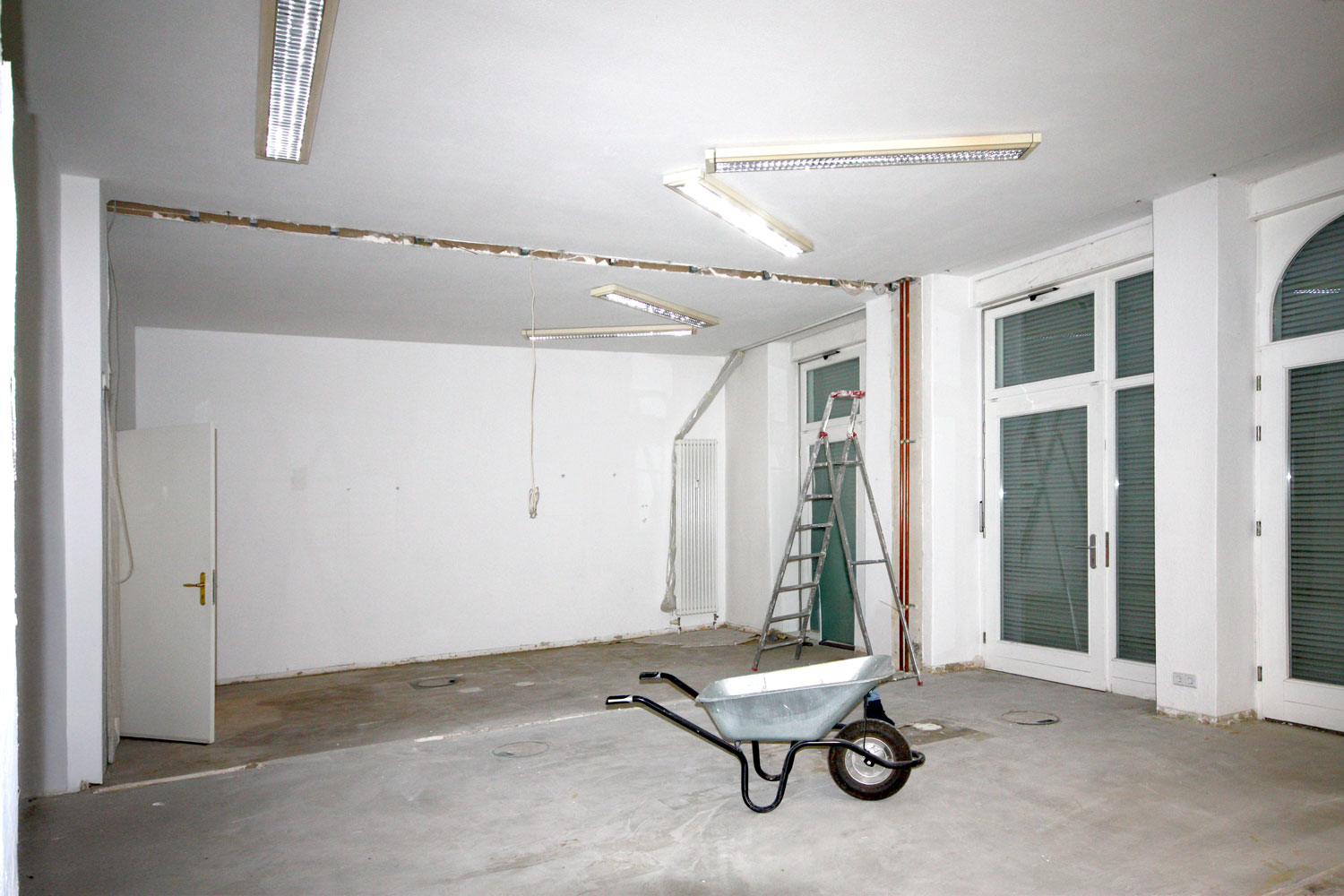 Vorherbild der Baustelle. Ein Raum mit Schubkarre und Leiter und sichtbarer Fuge an der Decke, wo eine Wand herausgerissen wurde.