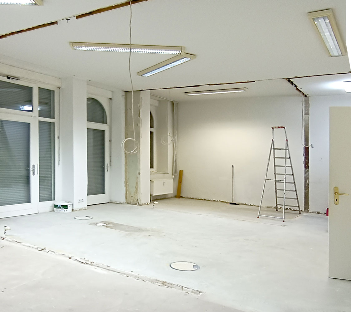 Vorherbild der Baustelle. Leerer Raum mit Leuchtstofflampen an der Decke und sichtbaren Fugen, wo Wände bereits abgerissen wurden.