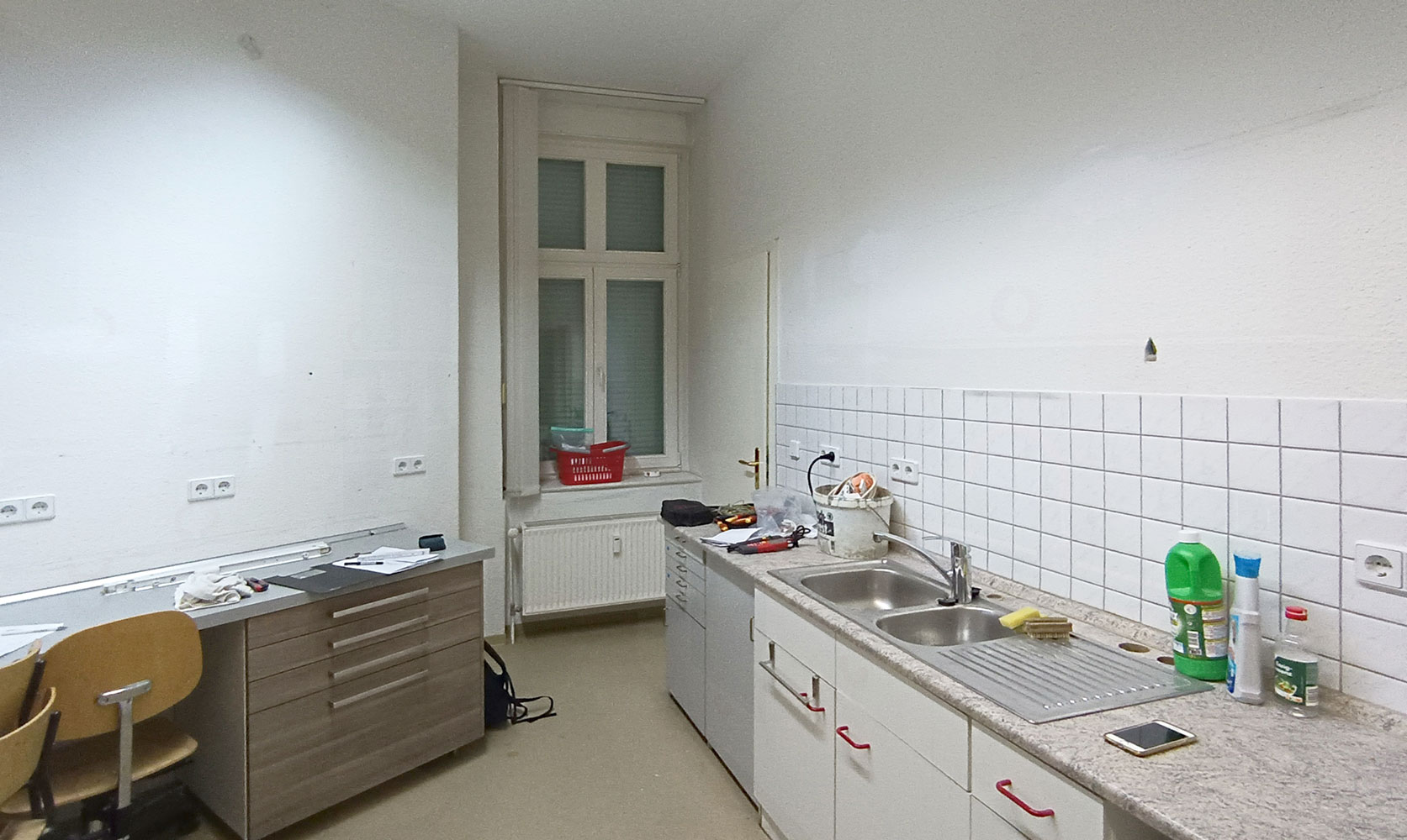 Vorher-Foto der Küche. Gebrauchte Küchenmöbel aus verschiedenen Materialien zusammengestellt. Weiße quadratische Fliesen als Spritzschutz, weiße Wände mit Raufasertapete. Beiger Linoleumboden.