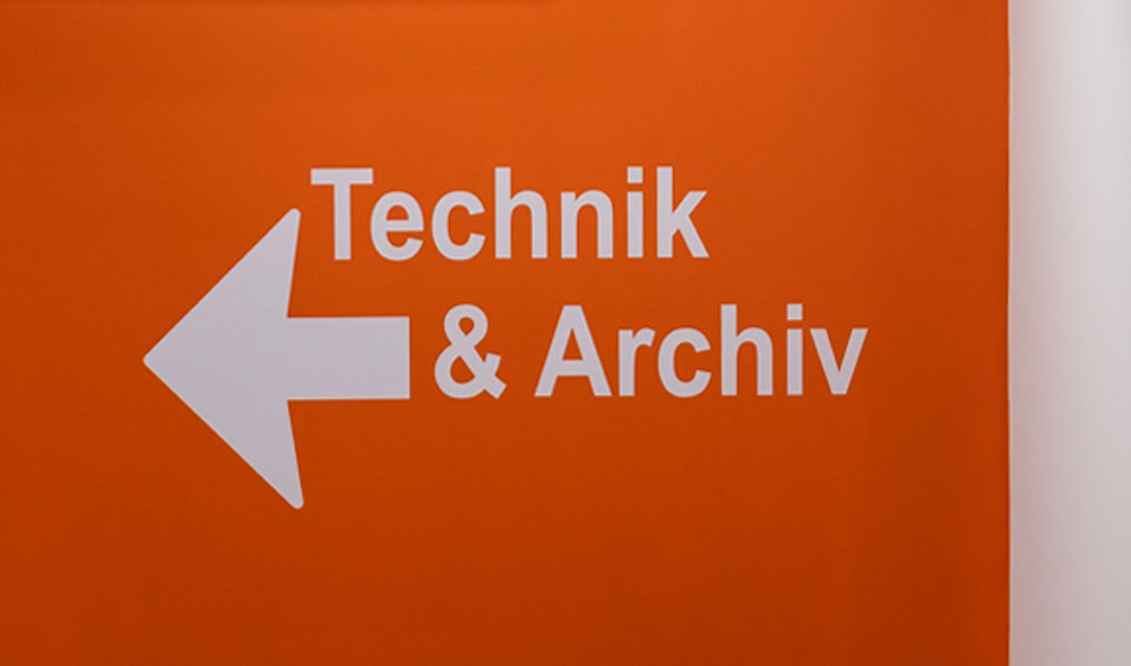 Orange Wand mit der Aufschrift Technik und Archiv und einem Pfeil. Es ist das Themenbild zu einem Beitrag zu Orientierung und Wegeleitung und der Begriffsbestimmung in diesem Themenfeld.