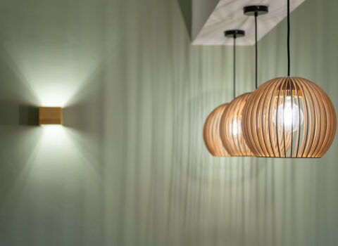 Lichtstimmung zum Wohlfühlen. Man sieht drei angeschaltete Pendelleuchten mit Holzlamellen und eine würfelförmige Wandleuchte aus Holz auf grünem Untergrund.