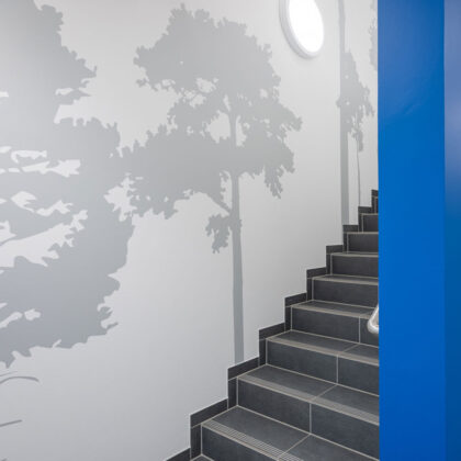 Ausschnitt eines Treppenaufgangs. Man sieht rechts ein Stück strahlend blaue Wand. In der Mitte Treppenstufen mit dunkelgrauen Fliesen und an der Wand graue Kiefernsilhouetten auf weißer Wand.