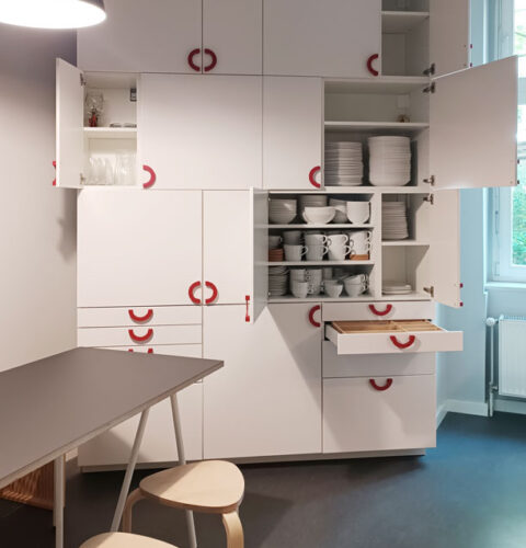 Küchenschrank aus weißen Möbeln, asymmetrisch angeordnet, mit roten Griffen, im Bild geöffnet. Man sieht das Geschirr für die gemeinschaftliche Nutzung.