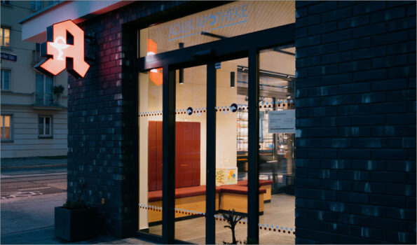 Eine Apotheke am Abend von außen mit einem leuchtenden Apothekenschild. Einblick in den Innenraummit Sitzbänken. Es ist das Beitragsbild zum Blogbeitrag "Apotheke barrierefrei".