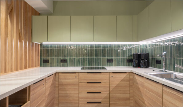 Wohlfühlen im Pausenraum: Das Bild zeigt eine Küchenzeile in einem Institut mit grüner Wand und grünen Oberschränken, glasierten grünen Fliesen und Eiche-Unterschränken sowie einen Raumteiler aus Eiche-Lamellen.