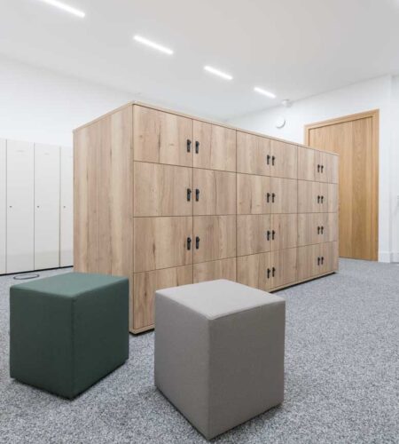 Lockerschränke in Eichedekoar und im Hintergrund weiße Spindschränke. Davor ein dunkelgrüner und ein hellgrauer würfelförmiger Polsterhocker.