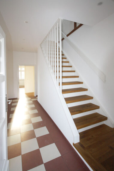 Flur in einem denkmalgeschützten Haus. Rot-weiße Fliesen und eine weiße Treppe mit Eichetrittstufen. Das Sonnenlicht scheint von hinten durch ein Zimmerfenster.