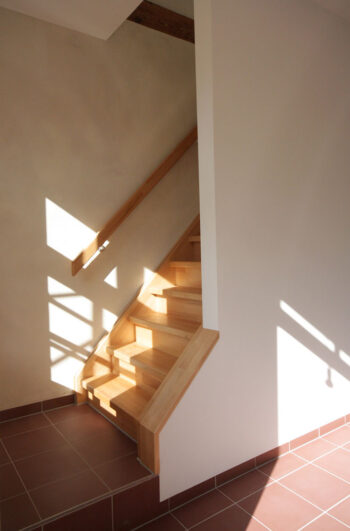 Stimmungsbild: Sonnenlicht scheint auf eine Buchenholztreppe. Der Boden und das Antrottspodest sind mit terrakotta-roten Fliesen belegt.
