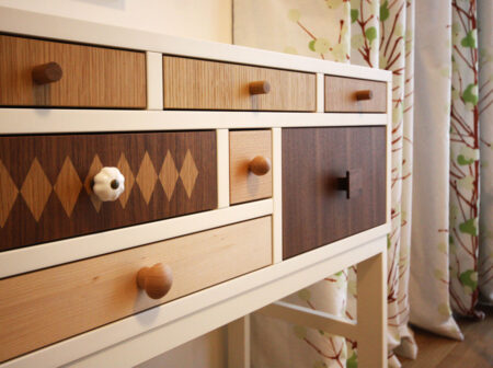 Sideboard in Weiß und mit verschiedenen Holzarten. Im Hintergrund ein Vorhang mit grünem Baummotiv.