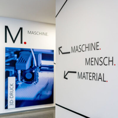 Nahaufnahme einer Wandgestaltung: Schriftzug Maschine. Mensch. Material." im Vordergrund. Im Hintergrund ein blaues Maschinenedetail eines 3D-Druckers.