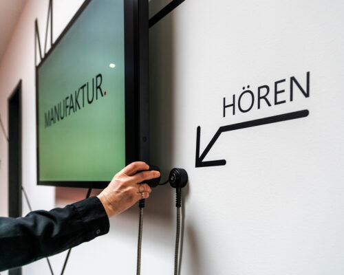 Nahaufnahme einer Hörstation neben einem Touchscreen. Auf der Wand sieht man das Wort "Hören" und einen Pfeil, der auf de Einhandhörer zeigt.