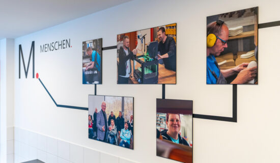 Teil einer Ausstellung: Zusammen mit dem Schriftzug "Menschen" sieht man rechteckige Fotos mit Mitarbeitenden bei der Arbeit.