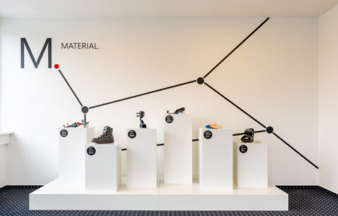 Blick auf eine weiße Wand mit schwarzen Linien und dem Schriftzug "Material". Davor steht ein weißes Podest und darauf 6 verschieden hohe weiße Säulen mit quadratischer Grundform. Auf den Säulen werden orthopädische Produkte als Ausstellung präsentiert.
