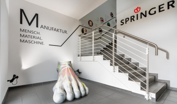 Ein kleines Foyer mit einem Treppenaufgang in einem pragmatischen Gewerbebau. Im Vordergrund eine überdimensionale Fußskulptur mit anatomischen Motiven als Projektion. Weiße Wände. An einer Wand das Logo der Springer Aktiv GmbH. An der anderen Wand die Schriftzüge "Manufaktur. Mensch. Maschine. Material". An der Wand des Treppenaufgangs ein Zeitstrahl zur Firmengeschichte mit kreisrunden Bildern und Text.