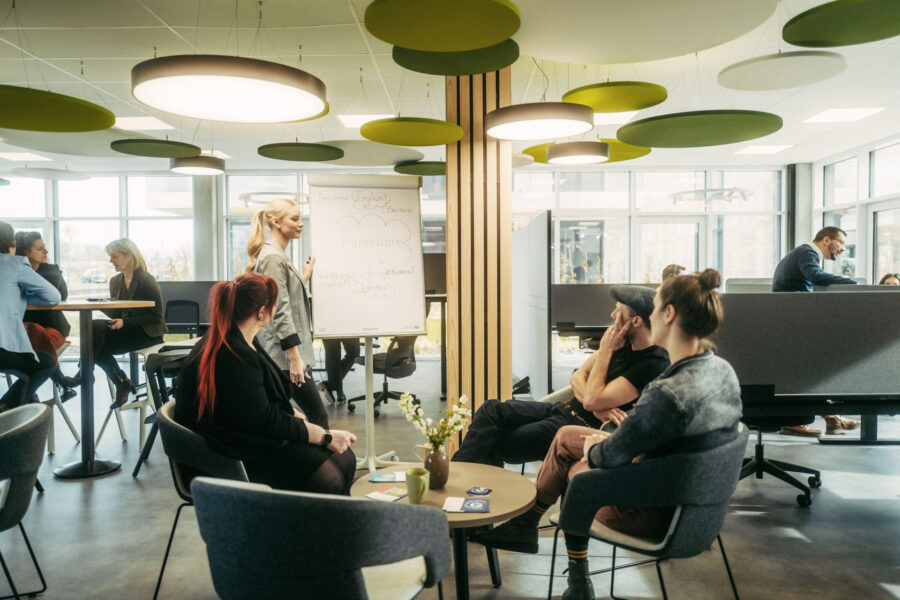 Ad-hoc-Besprechung an einem Flipchart in einer Coworking-Umgebung. Mittelpunkt des Raumes ist eine Säule mit Holzleisten und runden, weißen und grünen abgehängten Deckenabsorbern, die zusammen einen stilisierten Baum darstellen.