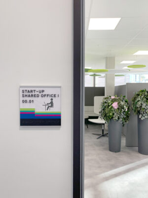 Wegeleitsystem mit farbigen Streifen auf einem Türschild; im Hintergrund ein Blick in einen Coworking-Space