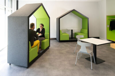 Ein Raum mit zwei schmalen, hausförmigen und rollbaren Sitzinseln. In der vorderen sitzen zwei Personen im Gespräch. Ein quadratischer Cafeteriatisch mit zwei Schalenstühlen ergänzt das Bild.