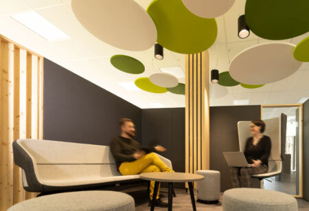 Eine Sitzgruppe in einer Coworking-Fläche. Ein mit hellen Holzlatten verkleideter Pfeiler und Deckensegel in Grüntönen und weiß bilden einen stilisierten Baum mit Blätterdach. Der Platz wird begrenzt durch halbhohe graue Wände. Das Mobiliar besteht aus Polsterhockern, Sofa und Hochlehner-Sessel. Zwei Personen sitzen sich im Gespräch gegenüber.