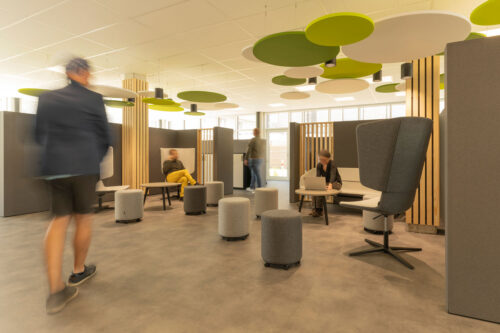 Ein zentraler Treffpunkt in einer Coworking-Fläche. Zwei mit hellen Holzlatten verkleidete Pfeiler und Deckensegel in Grüntönen und weiß bilden zwei stilisierte Bäume mit Blätterdach. Der Platz wird begrenzt durch halbhohe graue Wände. Das Mobiliar besteht aus Polsterhockern, Sofa und Hochlehner-Sesseln. Vier Personen beleben das Bild.