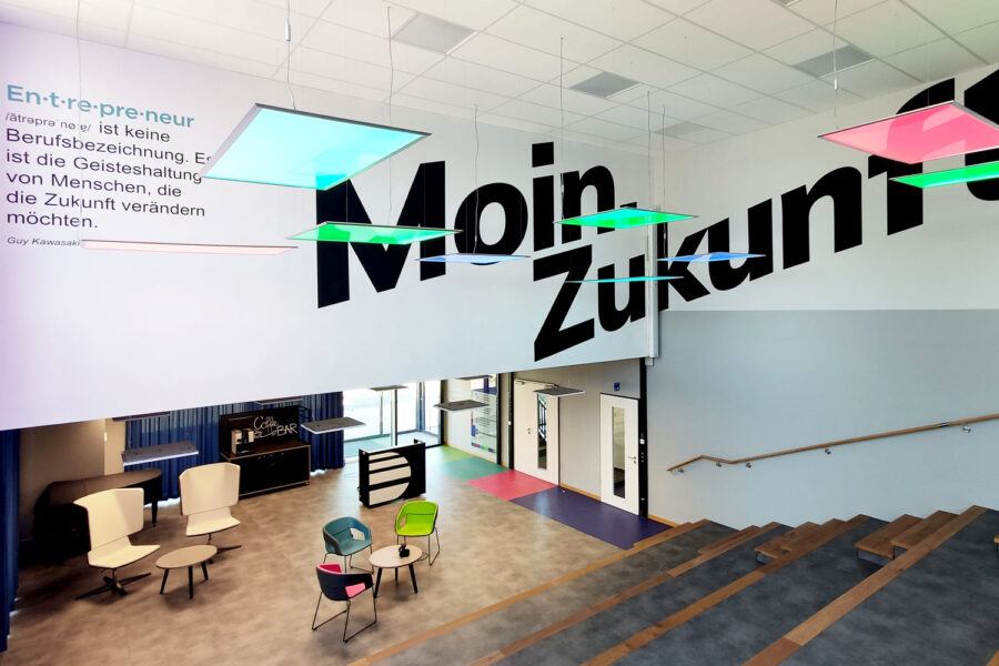 Das Foyer des Gründerzentrums DeveLUP in Ludwigslust. Ein Blick von einer großen Treppe mit Sitzstufen Richtung Eingang. Von der Decke hängen farbig leuchtende LED-Paneele. An der Wand ein großer schwarzer Schriftzug: Moin Zukunft sowie eine Definition des Begriffs Entrepreneur von Guy Kawasaki. Unten im Eingangsbereich sieht man Armlehntühle in den Corporate Design Farben hellgrün, cyan und magenta. Auch der Boden zeigt diese Farben als Streifen, der zu den weiteren Räumen führt.