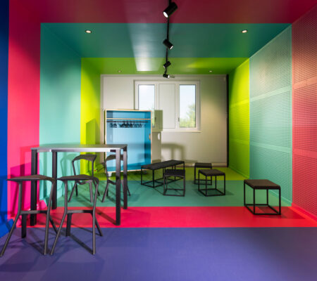 Ein Raum, bei dem Streifen aus den Corporate Design Farben hellgrün, magenta, cyan und ultramarin über Wände, Boden und Decke laufen. Zu sehen sind außerdem einige schwarze Möbel wie Sitzhocker, Stehtisch und Barhocker.