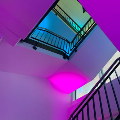 Treppenhaus mit schwarzem Geländer und weißen Wänden aus der Froschperspektive. Die Etagen sind für eine Abendveranstaltung mit farbigem Licht in den Corporate Design Farben inszeniert.