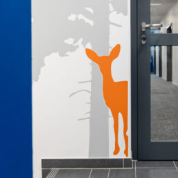 Dieses Kategoriebild führt zum Projekt: Flurgestaltung und Wegeleitung in der Autobahnmeisterei Michendorf. Es zeigt einen orangen Schattenriss eines Rehs vor einer Kiefernsilhouette als Wandbeklebung.