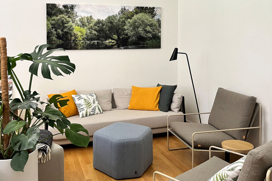 Polstersitzecke mit nachhaltigen Möbeln in Naturtönen. Ein Bild mit Bäumen an einer Fluss-Aue über dem Sofa. Links im Bild eine Pflanze. Als Farbtupfer gelbe Kissen auf dem Sofa.